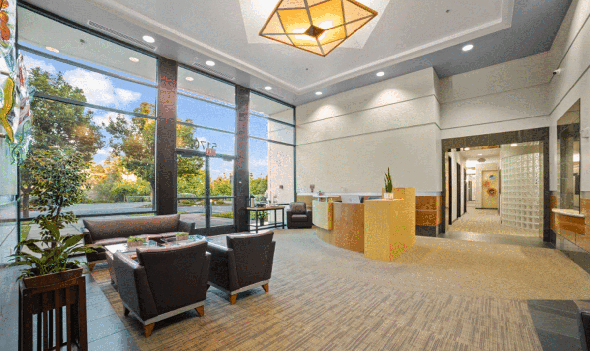 Bright office lobby with modern seating, reception desk, large windows, and views of trees outside